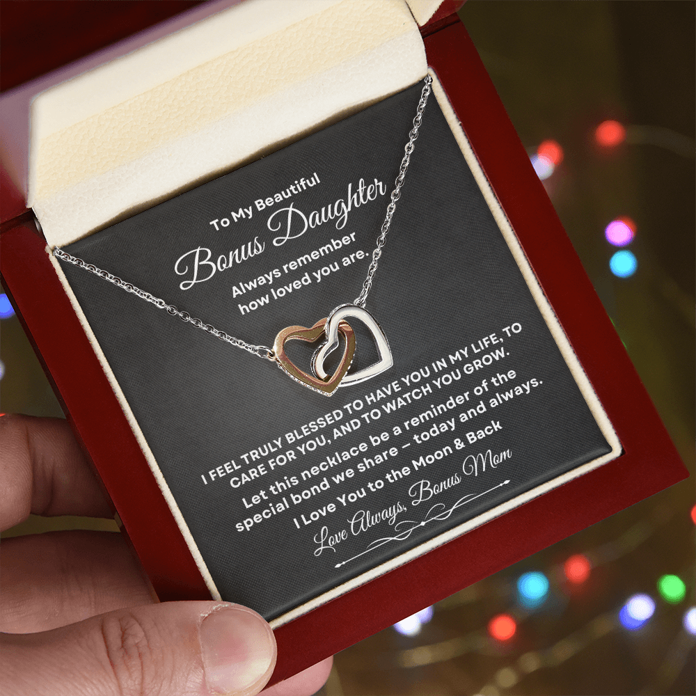 Hand holding an open mahogany luxury gift box with built-in LED light illuminating the interlocking hearts necklace and Bonus Daughter message card, with festive holiday lights blurred in the background.