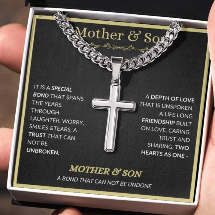 A stainless steel Cuban chain necklace with a cross pendant featuring a quote about the bond between a mother and son, presented in a black box with a quote about the necklace.