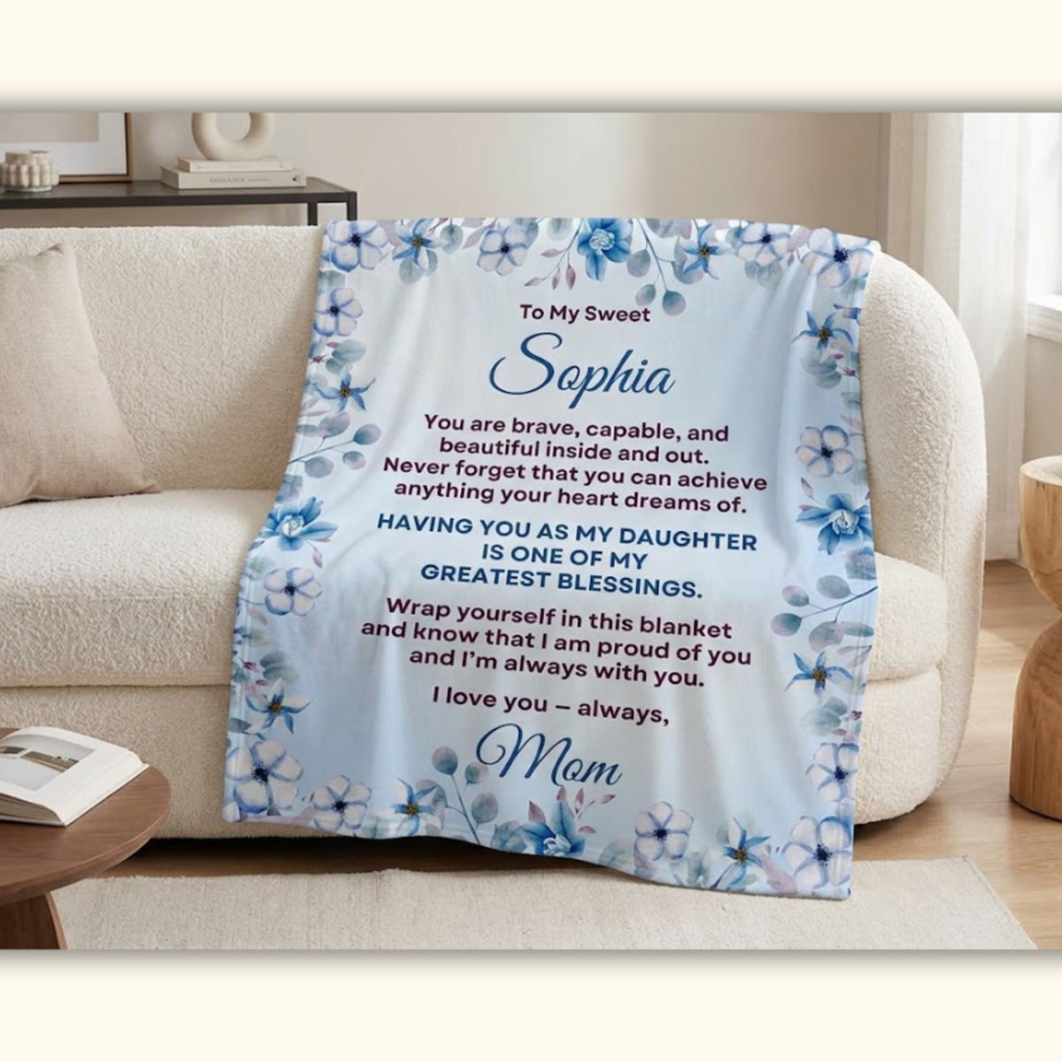 Personalized daughter blanket from Mom draped over a living room couch, featuring a cornflower blue floral design and heartfelt message for Sophia.