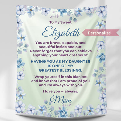 Personalized daughter blanket from Mom in soft sage with blue floral design, hanging against a white wall.