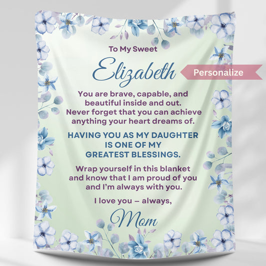 Personalized daughter blanket from Mom in soft sage with blue floral design, hanging against a white wall.